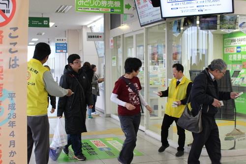 駅前で黄色いベストを着た二人の職員が啓発ティッシュを配布している様子の写真