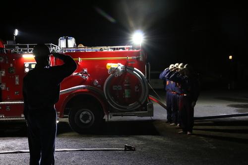 暗闇に光る消防車のライトに照らされる消防団員の皆さんの敬礼の写真