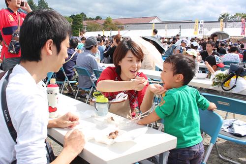 お母さんに大きな口を開けてご飯を食べさせてもらっている小さい子供と見守るお父さんの写真