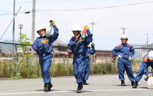 篠山市消防署内の訓練場を勇ましくホースを担いで行く2名の中学生と後ろから見守る消防隊員の写真