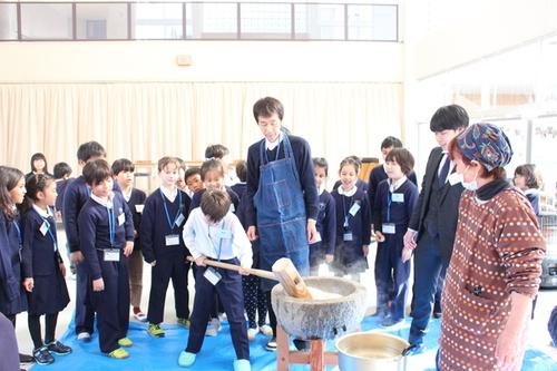杵を重そうに持つ男子生徒を見守る大人と楽しそうに見ている生徒達の餅つき体験の写真