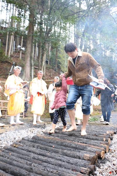 焼かれた黒い丸太の上を親子が手を繋ぎ裸足で歩く火渡りの儀式の写真