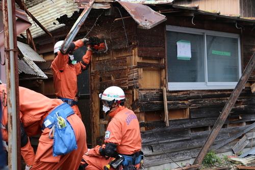 朱色の制服を身にまとった消防隊員の方達が協力して、エンジンカッターで木造の家屋の壁を切断している様子の写真