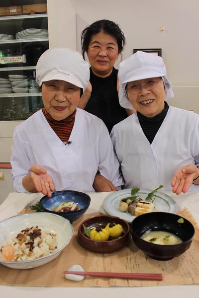 テーブルに並んでいる郷土料理を前に笑顔を浮かべている講師の森本さんと田中さんと2人の後ろにいるスタッフの西牧さんの写真