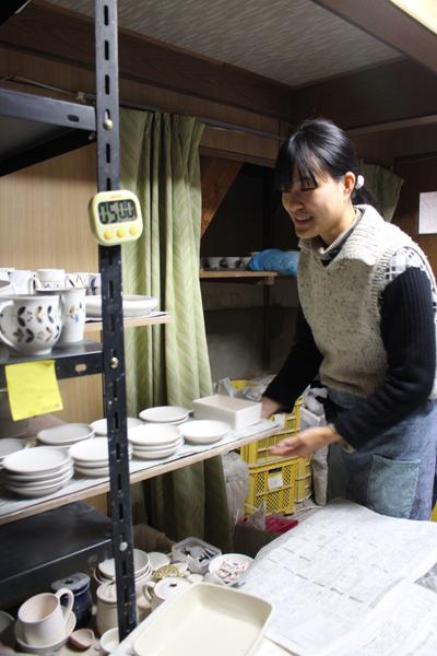 棚にたくさんの陶器がならぶ古民家を改修した工房で笑顔で陶器を整理する矢倉藍子さんの写真
