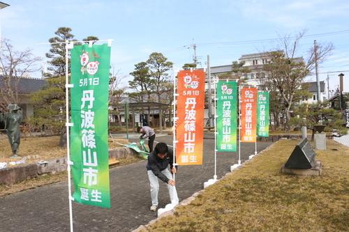 二人の男性がのぼり旗「5月1日丹波篠山市誕生」をたてる作業をしている様子の写真