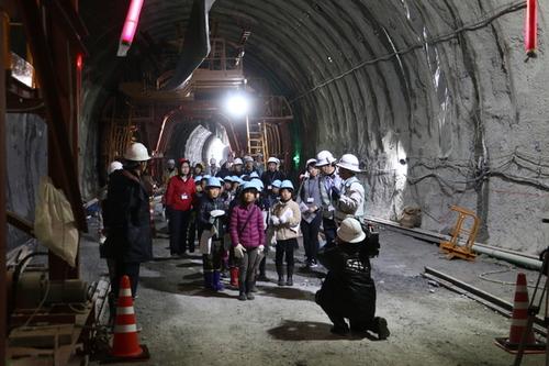 工事中のトンネル内でヘルメットをかぶり工事関係者の方から説明を受けている児童たちの写真