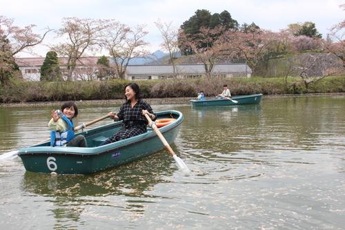 周囲に桜が咲く池の中で、救命胴衣を装着して嬉しそうに手を振る子供と笑顔で見守りながら観光ボートをこぐお母さんの写真