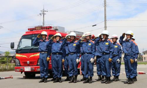 消防車の脇で整列し白いヘルメットを被り青い制服姿で敬礼する12名の中学生の写真