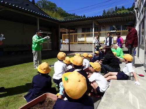 校舎の外で、男性講師の説明を、帽子をかぶり座って聞く生徒たちの写真