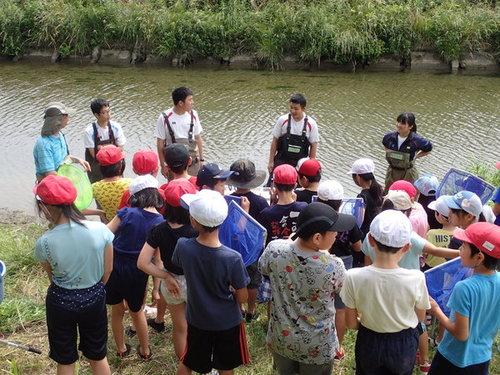 川を背にして説明を行う講師の皆さんと網を持って集まる城東小学校の生徒達の写真
