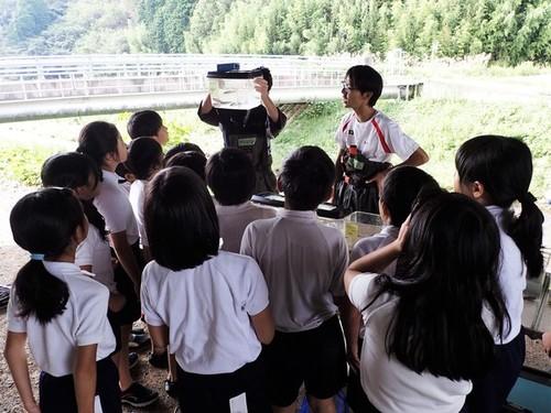 生き物が入った水槽を持ち上げて説明する田井先生と生徒の皆さんを興味深そうに見つめる小学生の皆さんの写真