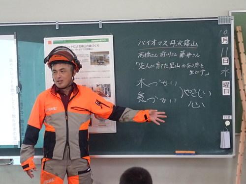 オレンジとグレーの色が入った作業着にヘルメットを着用した講師の方が、バイオマス丹波篠山について黒板を使って説明している写真