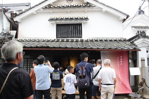 ピンク色の暖簾が玄関前にある白壁瓦屋根の江戸商家風家屋とその前に集まって中を見たり写真を撮る人々が多く集まった様子の写真