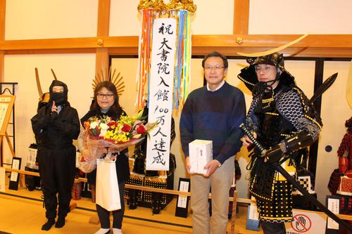 甲冑と忍者姿の男女2人と花束と贈呈品を持った男女がくす玉の前に並んだ篠山城大書院100万人達成祝いの写真