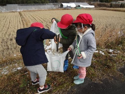 協力してゴミ袋を持ちトングを使ってゴミを入れようとしている3人の園児の写真