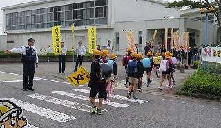 警察官の方が右手を上げて見守る横断歩道を黄色い旗を持った児童たちが1列になってわたる写真