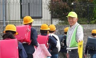 1列になって登校する児童たちを黄色い旗を持って笑顔で見守る1人の男性の写真