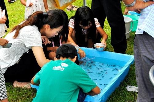 芝生に置かれた水色の水槽を囲んで夢中で金魚すくいを楽しむ子供たちの写真