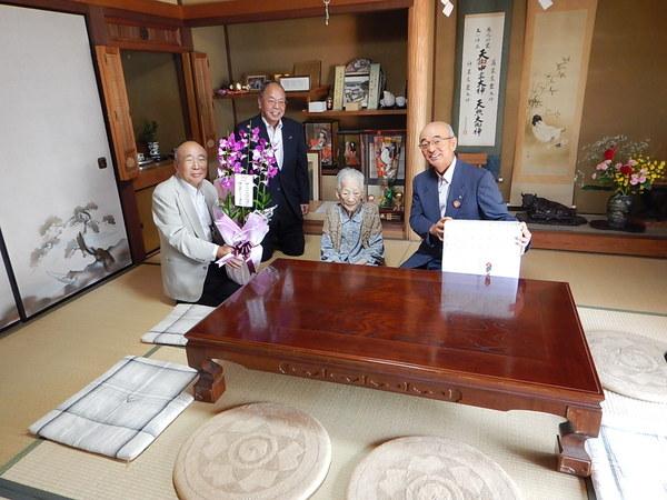 床の間のテーブルの前で記念品と花に囲まれて座っている岸本 冨子さんが市長らと一緒に記念撮影している写真