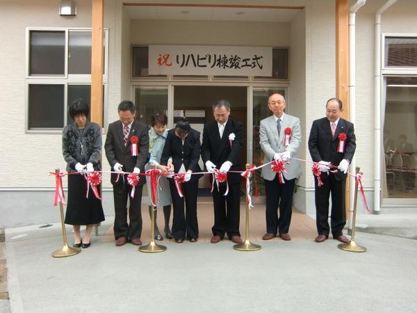 市長と一緒に5名の人達が横一列に並び、祝リハビリ棟竣工式と書かれた幕の前でテープカットをしている写真