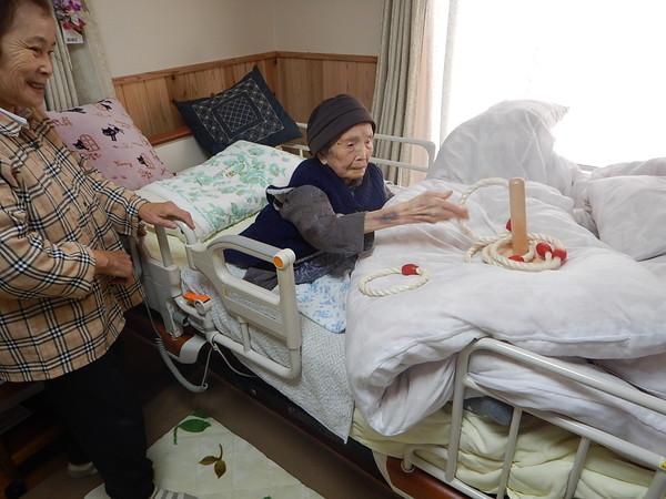 帽子を被った塚西 す江さんがベッドの上で輪投げをしている様子を横で笑顔で見ている女性の写真