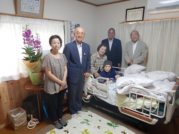 ベッドに座っている帽子を被った塚西 す江さんが花や市長らと一緒に記念撮影している写真