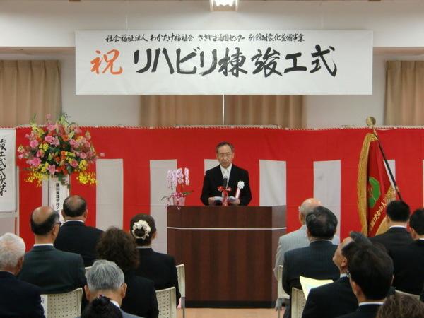 祝リハビリ棟竣工式と書かれた幕の前で、胸に花をつけてスーツを来た理事長が参加者へ話をしている写真