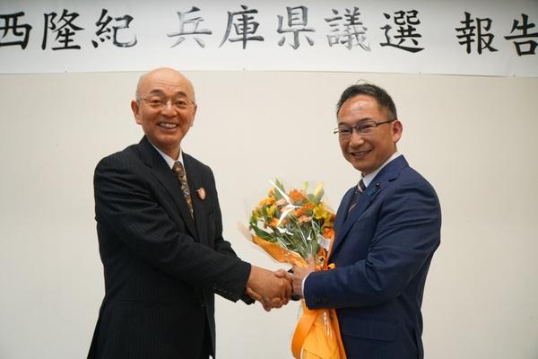小西 たかのりさんが花束を手に持ち市長と笑顔で握手をしている写真