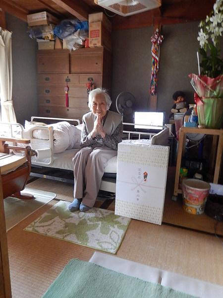 自宅のベッドで手を合わせて座っている野垣 こちよさんが花や記念品と一緒に記念撮影している写真