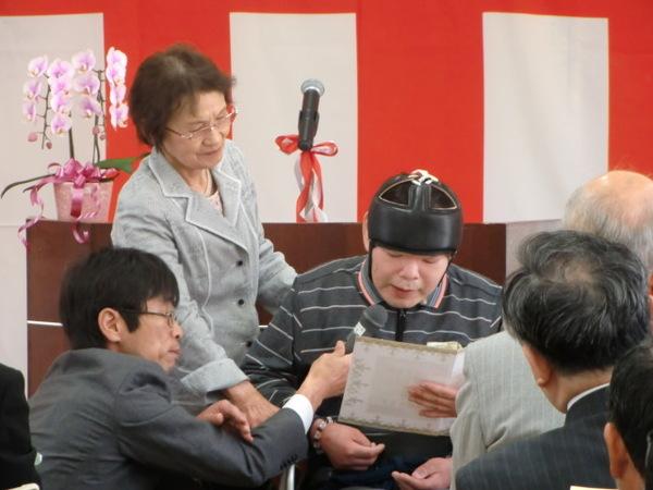 頭にヘッドギアを付けた、リハビリ棟の利用者の方が付き添いの男性にマイクを持ってもらって話をしている写真