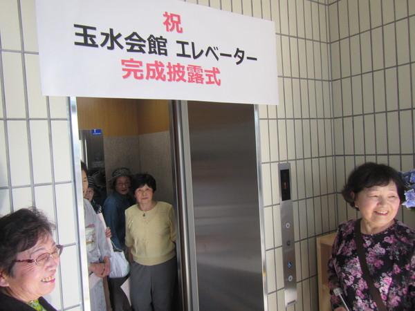 「祝 玉水会館にエレベーター完成披露式」の横断幕のかかっているエレベータに実際に女性が数名乗って利用しようとしており、エレベーターの入り口にいる女性が笑顔で写っている写真