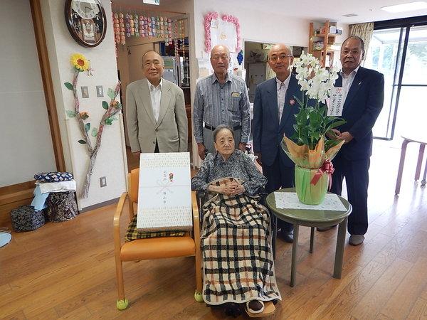 車椅子に座っている酒井 しず子さんが記念品と花に囲まれて市長らと一緒に記念撮影している写真