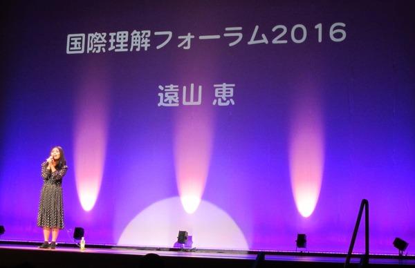 ステージに赤紫のライトが照らされ、遠山 恵さんが歌っている写真