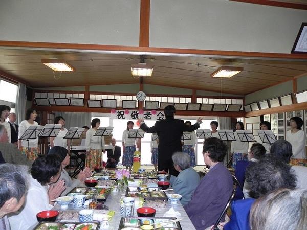 敬老会で女性の方達が歌を披露して、年配の方が聞きながら手拍子をしている写真