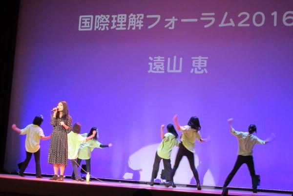 遠山 恵さんがステージで歌い、その後ろで6名の子供たちが歌に合わせてダンスをしている写真