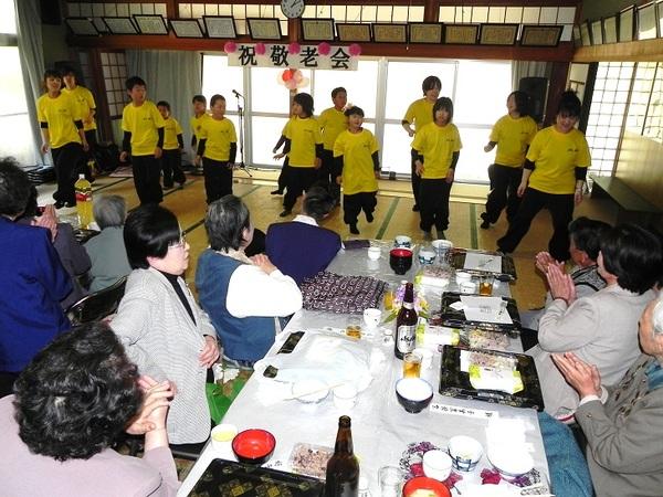 黄色い服を着た子供達がおじいちゃん、おばあちゃんに踊りを披露している写真