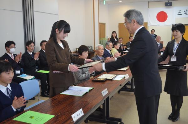 茶色の洋服を着た髪の長い女性が賞状を受け取っている写真