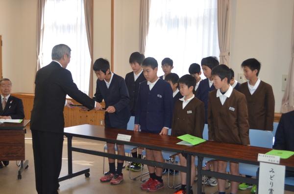 紺色の制服と茶色の制服を着た子供達がみんなで起立して代表の子供が賞状を受け取っている写真