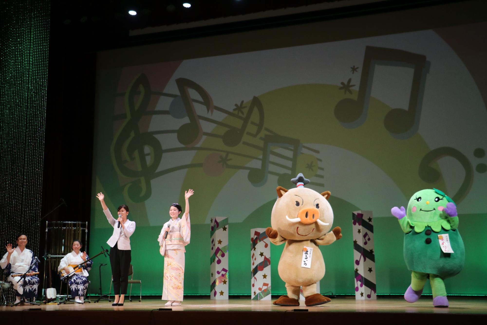 デカンショのど自慢の舞台で、司会者、チャンピオン、まるいの、まめりんが手を振っている