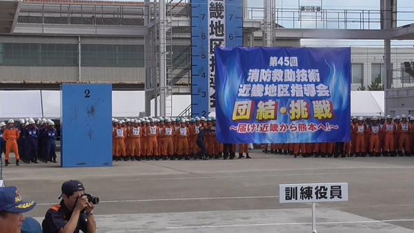 消防救助技術近畿地区指導会の大きな横断幕を飾ってあり、その後ろに大会へ参加している消防団員が整列している写真