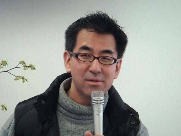 グレーのハイネックに黒いベストを着ており、マイクを持って話をしている松山 直樹さんの写真