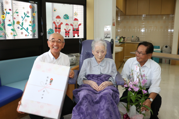 車いすに座っている岸本冨子さん(106歳)が記念品を持った市長らと一緒に記念撮影している写真