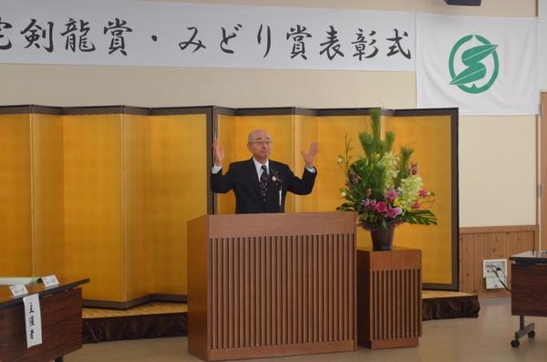 表彰式の壇上で綺麗な花ががざられた横で市長が両手をあげてたっている写真