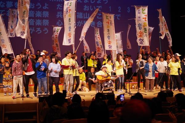 ホールの舞台にのぼり旗とスタンドマイクがたくさん立ててあり車いすに乗った男性が演奏するギターに合わせて歌っている人々の写真