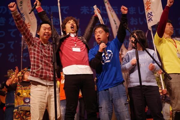 ホールの舞台にてマイクを持った男性らが手を上げて声を出している写真