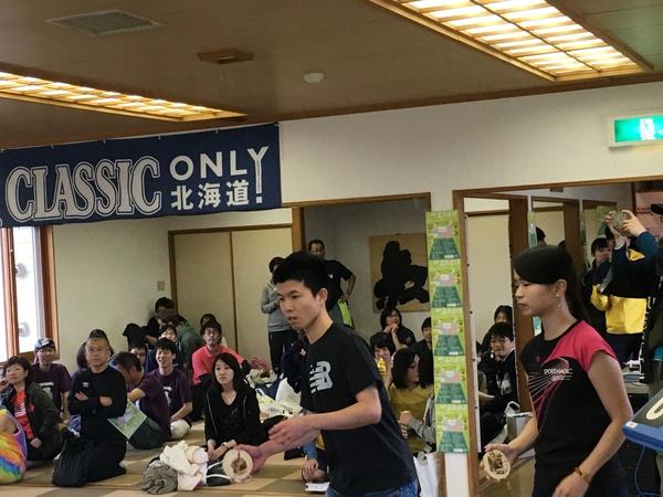 ユニホーム姿の男女ペアがラケットを持って構えているのを畳に座って人々が見ている写真