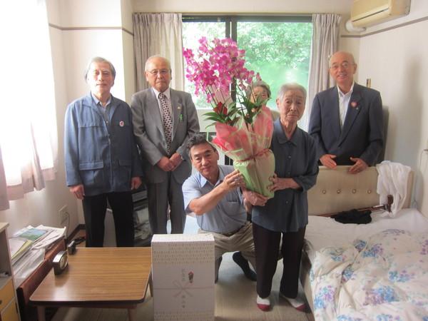 介護施設にて長寿祝いの花を持った松本まつヱさん(101歳)と市長らとの記念撮影している写真