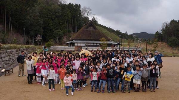 公園に集まったたくさんの子供達がピースサインをして後ろで大人の人達が手をあげている集合写真