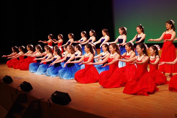 ホールの舞台で髪飾りを付け赤色や青色の衣装を着た女性らが前列は座り、後列は立って踊っている写真